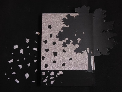 silueta de un árbol sobre un papel plateado brillante. Pedacitos de papel negros simulan ser las hojas del árbol. Cuando el papel brillante se acaba, las hojas negras pasan a ser parte de un fondo oscuro y se vuelven plateadas brillantes.